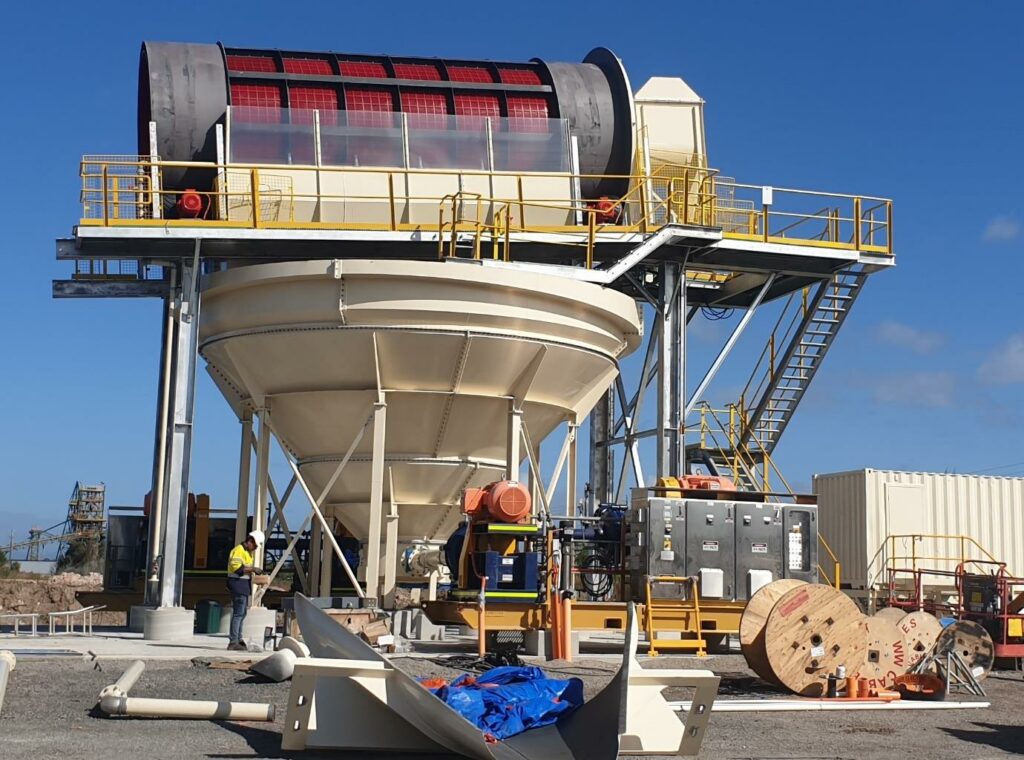 An industrial facility with a large cylindrical structure supported by a metal frame, featuring a funnel-shaped base, red panels, and additional machinery including a Trash Screen. A worker is present near the structure.